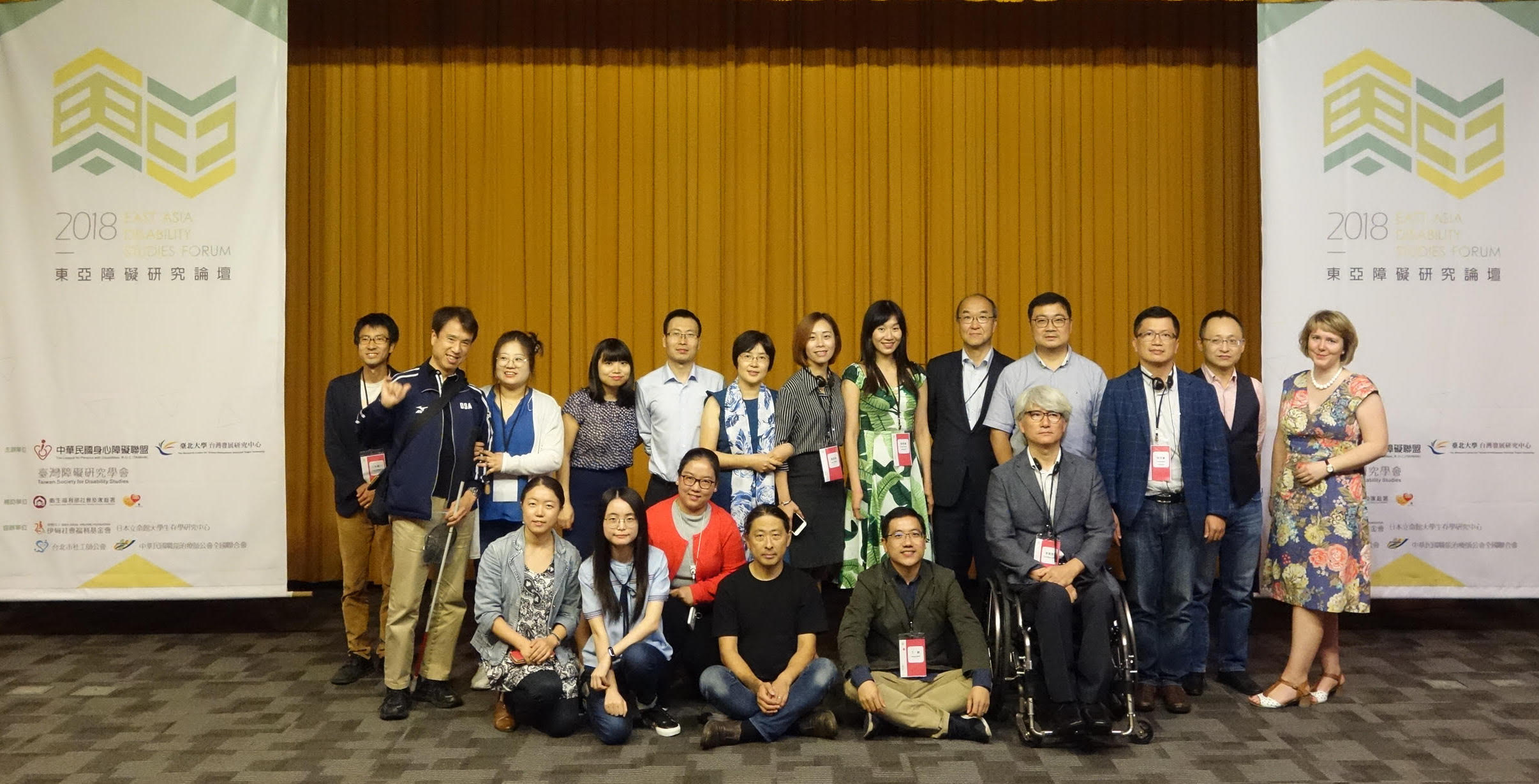 這張照片有19位來自各國的與會者，兩側為一層樓高的論壇海報