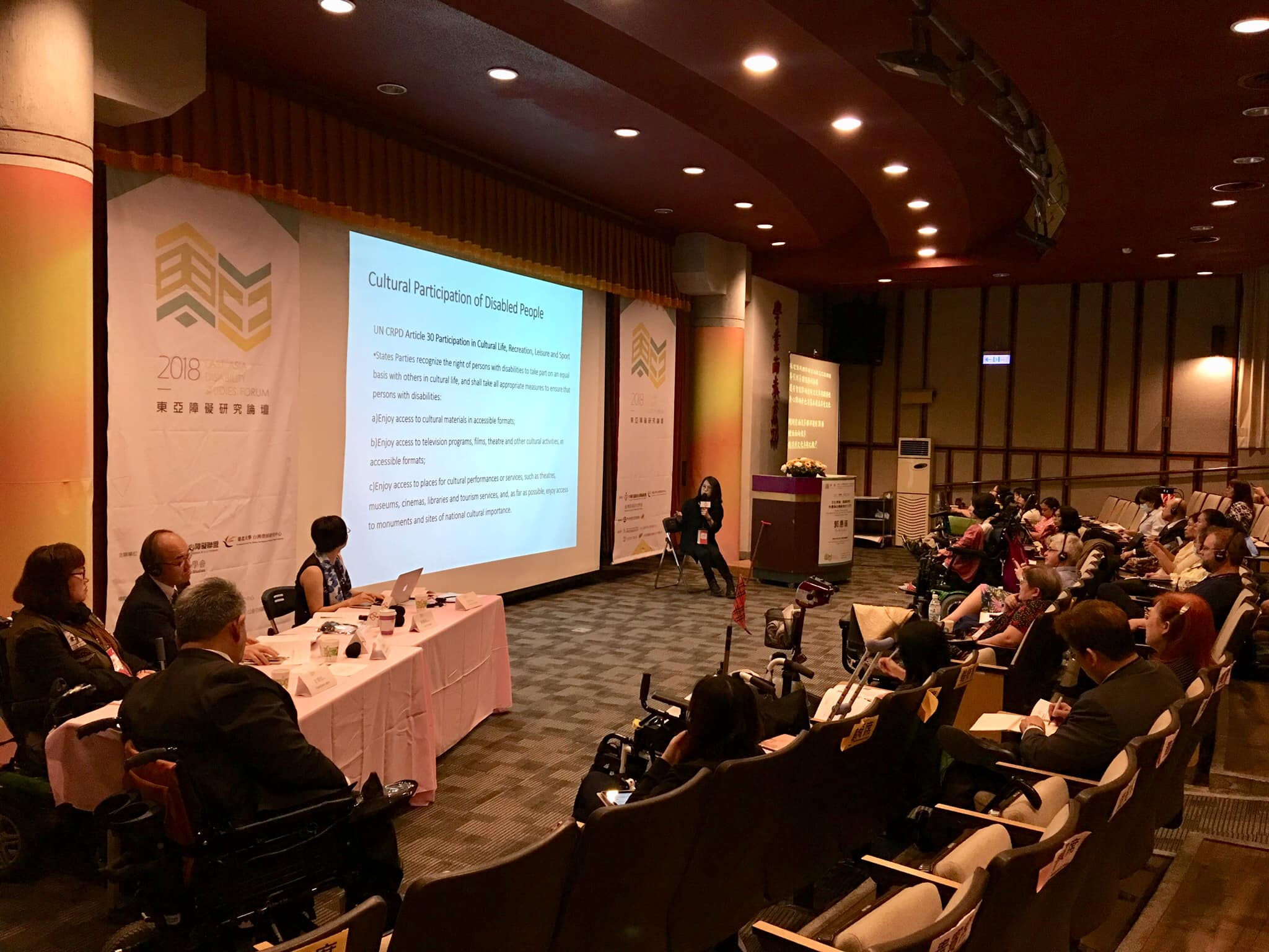 論壇發表講者郭慧瑜身著黑衣坐在大螢幕前報告研究，可見講堂一排排觀眾與前台角落的數位發表人